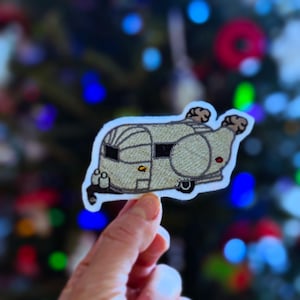 May include: Embroidered patch of a vintage travel trailer in shades of tan and brown, with black windows and a red accent. The patch has a white border and is held against a blurred background of Christmas tree lights.