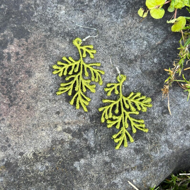 May include: A pair of lime green leaf-shaped earrings with silver-colored hooks. The earrings are made of a textured material and are displayed on a gray stone surface. The earrings have a detailed, naturalistic design, resembling a sprig of foliage.