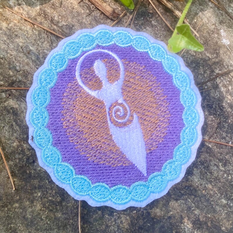 May include: Circular embroidered patch with a light purple figure on a purple and gold background, framed by a scalloped light blue border. The design is likely for application on clothing or accessories.