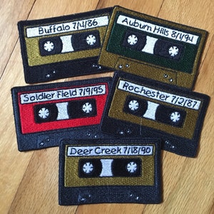 May include: Five embroidered patches shaped like cassette tapes. Each patch has a different color and text: "Buffalo 7/4/86", "Auburn Hills 8/1/94", "Soldier Field 7/9/95", "Rochester 7/2/87", and "Deer Creek 7/18/90". The patches are on a wooden surface.