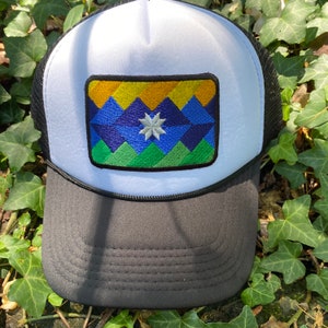 May include: A trucker hat with a white front panel and a black mesh back. The hat features a rectangular patch with a geometric mountain design in shades of yellow, blue, and green, and a white star. The brim is black.