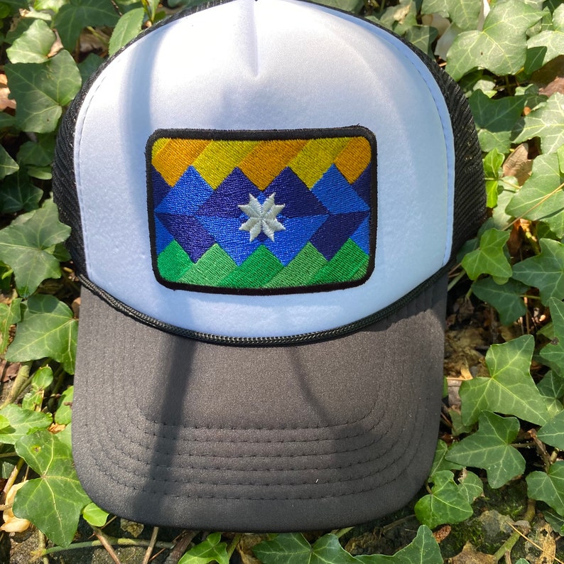 May include: A trucker hat with a white front panel and a black mesh back. The hat features a rectangular patch with a geometric mountain design in shades of yellow, blue, and green, and a white star. The brim is black.