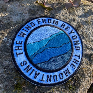 May include: A circular embroidered patch with the text "THE WIND FROM BEYOND THE MOUNTAINS" in a blue arc. The central design shows layered blue mountain shapes within a blue circle, creating a landscape.