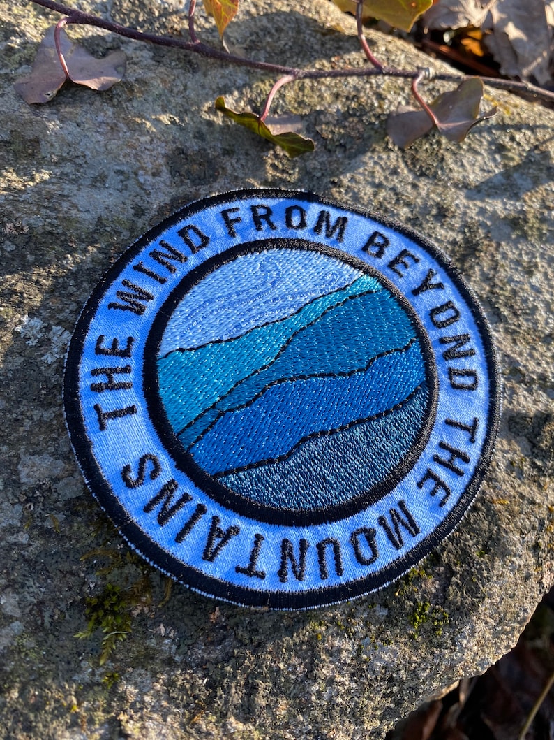 May include: A circular embroidered patch with the text "THE WIND FROM BEYOND THE MOUNTAINS" in a blue arc. The central design shows layered blue mountain shapes within a blue circle, creating a landscape.