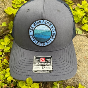 May include: Gray trucker hat with a dark blue mesh back and a circular patch featuring a mountain landscape and the text "THE WIND FROM BEYOND THE MOUNTAINS". The hat has a curved brim and an adjustable strap.