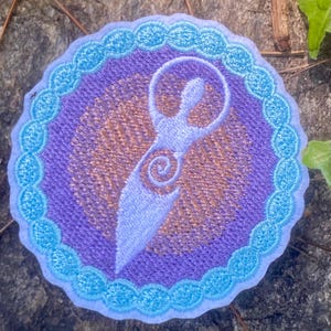May include: Embroidered patch with a light blue figure in a circle. The figure has a spiral design, set against a purple background with a gold pattern. The patch is bordered by a light blue scalloped edge, suitable for clothing or accessories.