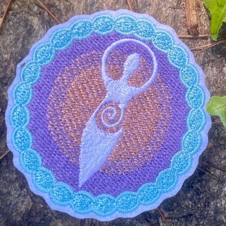 May include: Embroidered patch with a light blue figure in a circle. The figure has a spiral design, set against a purple background with a gold pattern. The patch is bordered by a light blue scalloped edge, suitable for clothing or accessories.