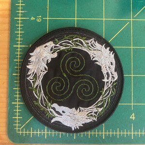 May include: Round black embroidered patch featuring three stylized white wolf heads surrounding a green triple spiral design. The wolves are embroidered in white thread, with green vines and leaves. The patch is approximately 4 inches in diameter.