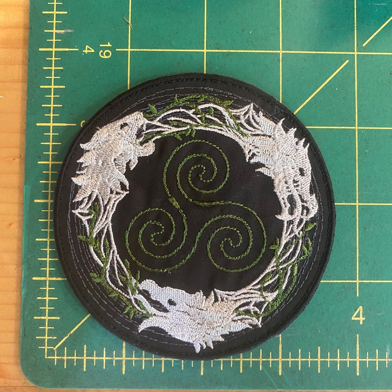 May include: Round black embroidered patch featuring three stylized white wolf heads surrounding a green triple spiral design. The wolves are embroidered in white thread, with green vines and leaves. The patch is approximately 4 inches in diameter.