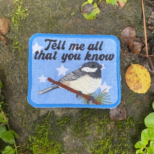 May include: Embroidered patch with a blue background and white stars. The patch features a detailed illustration of a small bird perched on a branch, with the text "Tell me all that you know" in black script.