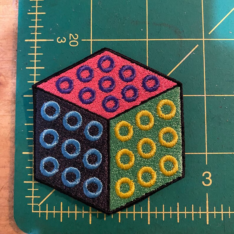 Puede incluir: Parche bordado con un dise&ntilde;o de cubo geom&eacute;trico. El cubo est&aacute; dividido en cuatro secciones: negro, rosa, verde y negro. Cada secci&oacute;n presenta c&iacute;rculos bordados en azul, morado y amarillo. El parche tiene un borde negro.