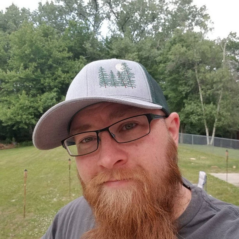 May include: A gray trucker hat with a forest and moon design. The hat has a gray front panel and a dark green mesh back. The design features a crescent moon and a cluster of pine trees in green. The hat is worn by a person with a red beard and glasses.