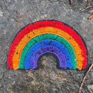 May include: A colorful rainbow patch with red, orange, yellow, green, blue, and purple fringe. The rainbow is set against a dark background, creating a vibrant contrast. This decorative patch can be used to embellish clothing or accessories.