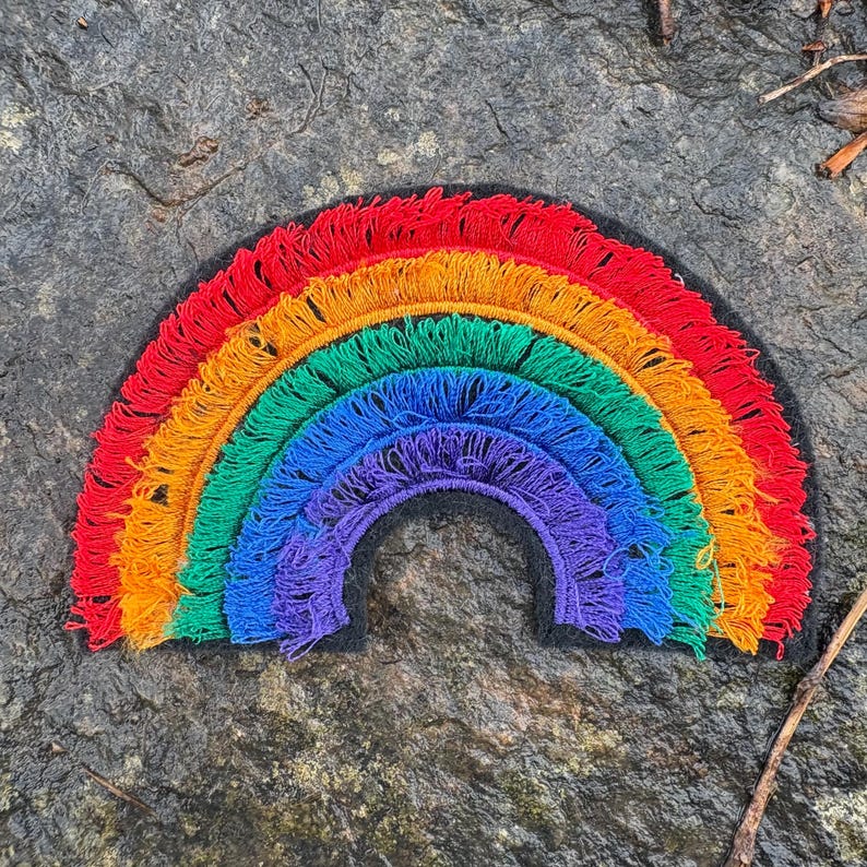 May include: A colorful rainbow patch with red, orange, yellow, green, blue, and purple fringe. The rainbow is set against a dark background, creating a vibrant contrast. This decorative patch can be used to embellish clothing or accessories.