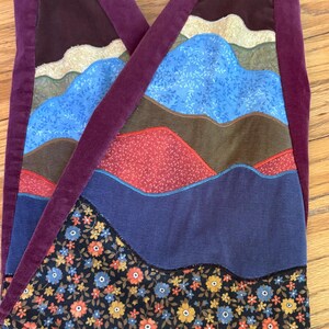 Op de afbeelding: Corduroy broek met een patchwork ontwerp met bergachtige vormen in verschillende kleuren, waaronder blauw, bruin, rood en bloempatronen. De broek heeft een dieppaarse corduroy tailleband en bandjes.
