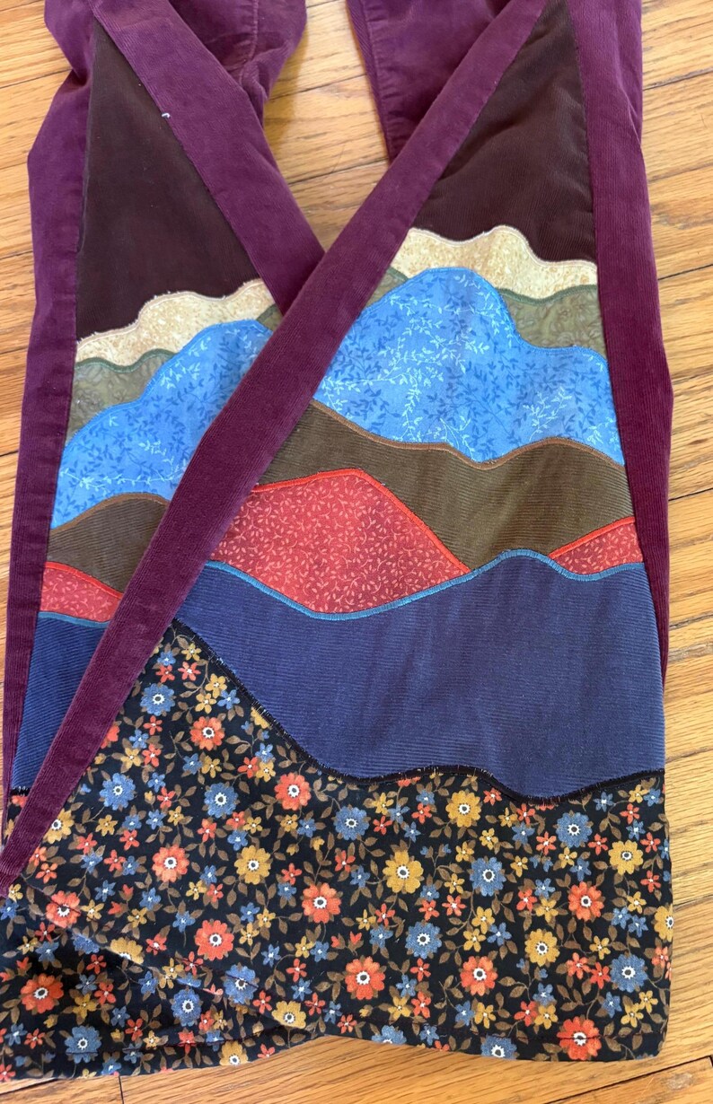 Op de afbeelding: Corduroy broek met een patchwork ontwerp met bergachtige vormen in verschillende kleuren, waaronder blauw, bruin, rood en bloempatronen. De broek heeft een dieppaarse corduroy tailleband en bandjes.