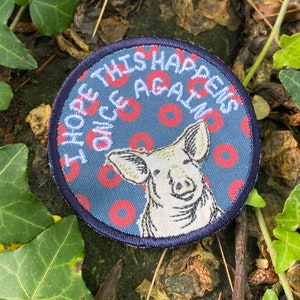 May include: Round patch with a pig illustration and the text "I HOPE THIS HAPPENS ONCE AGAIN". The patch has a blue background with red donut shapes and a navy blue border. The pig is white with embroidered details.
