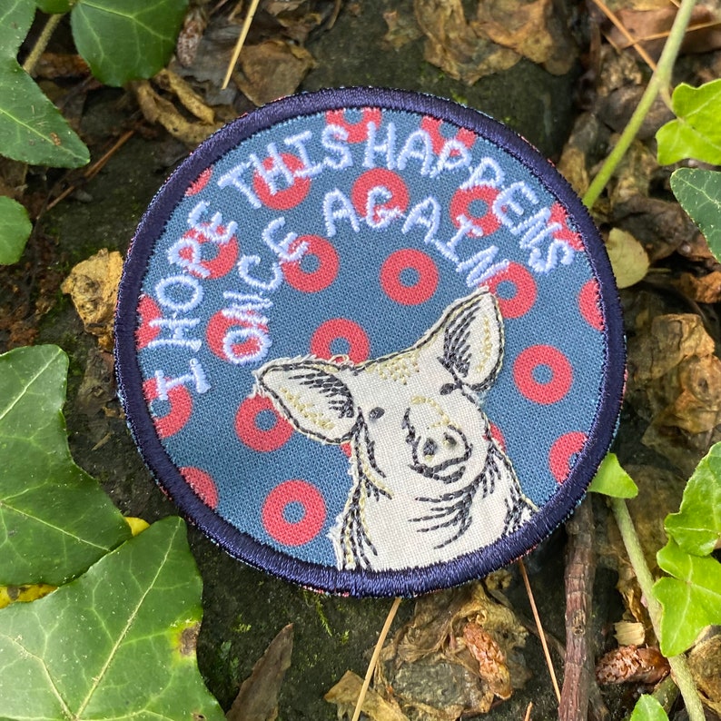 May include: Round patch with a pig illustration and the text "I HOPE THIS HAPPENS ONCE AGAIN". The patch has a blue background with red donut shapes and a navy blue border. The pig is white with embroidered details.