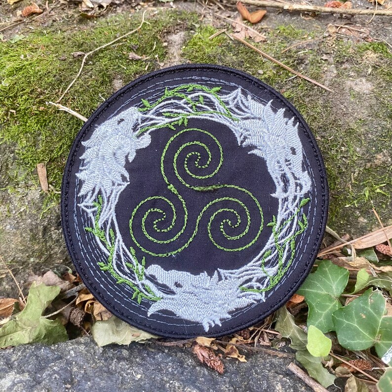 May include: Round black patch with a detailed embroidered design. The patch features three stylized wolf heads surrounding a green spiral symbol, with green vines and leaves. The wolves are light gray, and the stitching is precise.