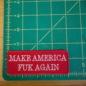 Może przedstawiać: Prostokątna czerwona naszywka z białym haftowanym napisem "MAKE AMERICA FUK AGAIN". Naszywka znajduje się na zielonej macie do cięcia z liniami siatki i wymiarami w centymetrach.