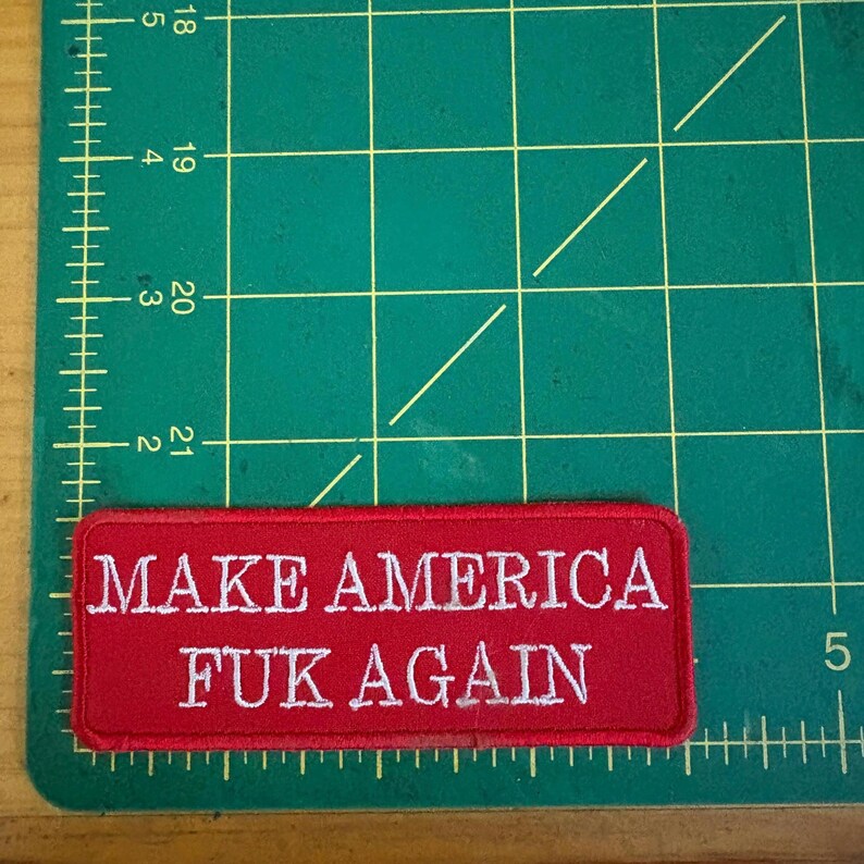Może przedstawiać: Prostokątna czerwona naszywka z białym haftowanym napisem "MAKE AMERICA FUK AGAIN". Naszywka znajduje się na zielonej macie do cięcia z liniami siatki i wymiarami w centymetrach.