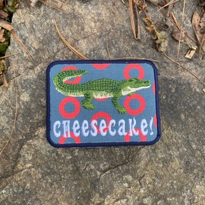 May include: Rectangular patch with a blue background featuring a green alligator and red circles. The word "CHEESECAKE!" is embroidered in white. The patch has a navy blue border.