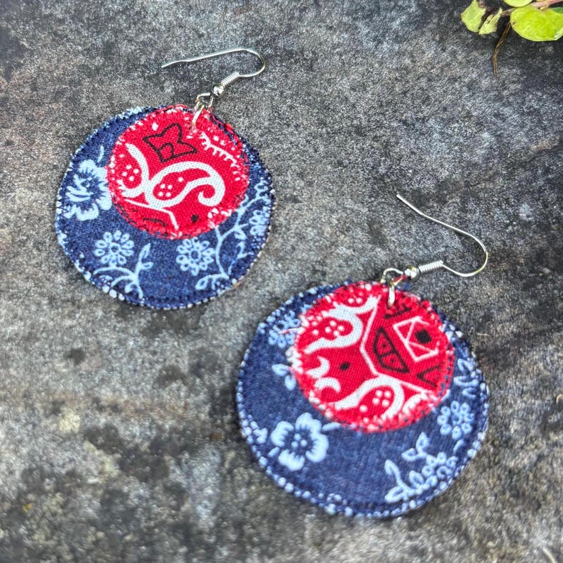 May include: A pair of handmade fabric earrings. Each earring features a red paisley center, surrounded by a navy blue floral patterned fabric. The earrings have silver-colored ear wires.