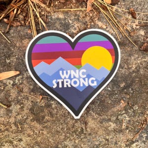 Puede incluir: Una pegatina en forma de coraz&oacute;n con un dise&ntilde;o de monta&ntilde;a y puesta de sol. La pegatina presenta una cordillera azul, un sol amarillo y el texto "WNC STRONG" en blanco. El fondo incluye tonos de morado, verde y rojo.