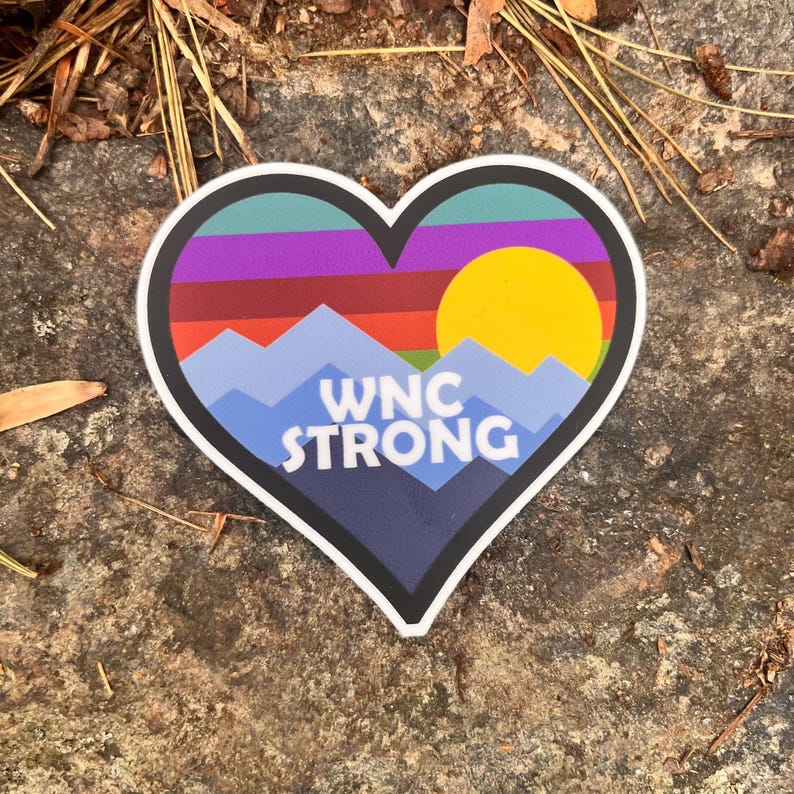 Puede incluir: Una pegatina en forma de coraz&oacute;n con un dise&ntilde;o de monta&ntilde;a y puesta de sol. La pegatina presenta una cordillera azul, un sol amarillo y el texto "WNC STRONG" en blanco. El fondo incluye tonos de morado, verde y rojo.