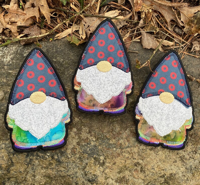 May include: Three gnome-shaped patches with a whimsical design. Each patch features a blue hat with red circles, a white beard, and a colorful, tie-dye-like body. The patches are outlined in black.