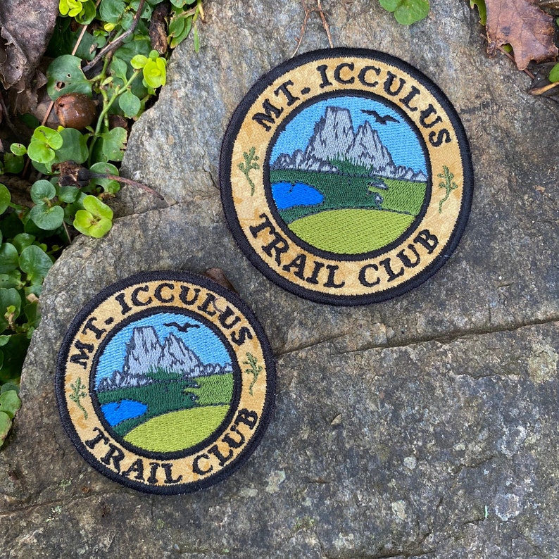 Op de afbeelding: Twee ronde geborduurde patches met de tekst "MT. ICCULUS TRAIL CLUB". De patches tonen een berglandschap met een blauwe lucht, een meer en groen gras. De rand is beige met zwarte rand.