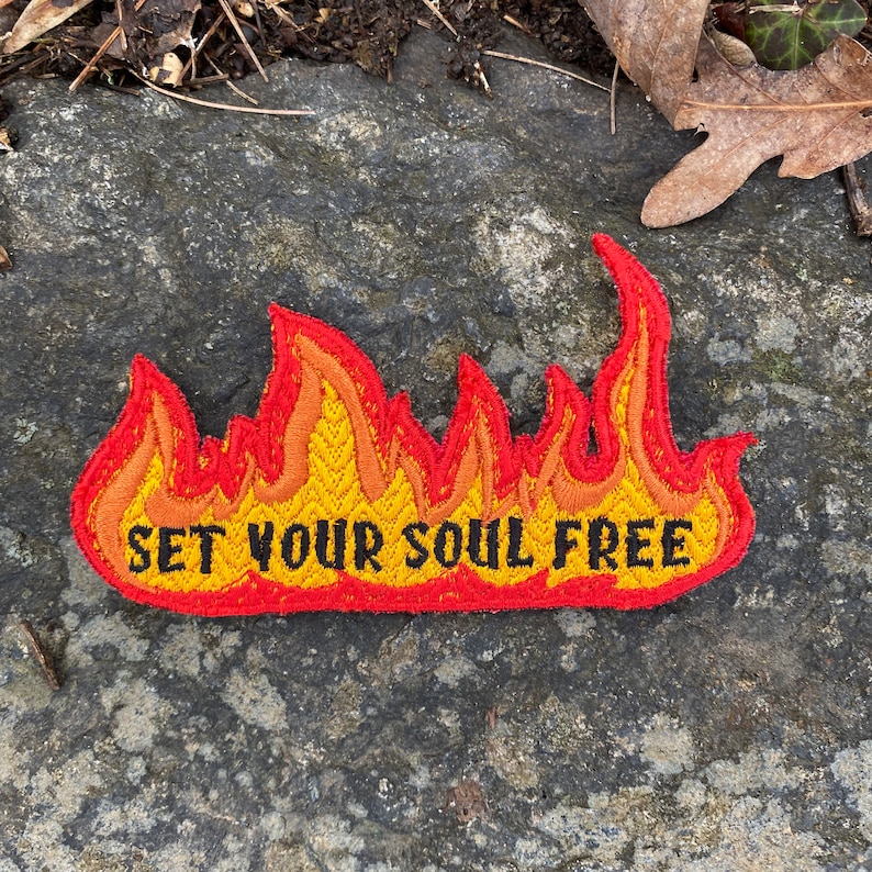 May include: Embroidered patch in the shape of flames, with a red border and yellow, orange, and red interior. The text "SET YOUR SOUL FREE" is embroidered in black across the centre of the flames.