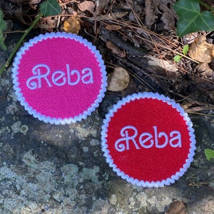 May include: Two circular embroidered patches. One is hot pink, the other is red. Both have the name "Reba" in white script. Each patch has a white scalloped border. The patches are on a gray stone surface.