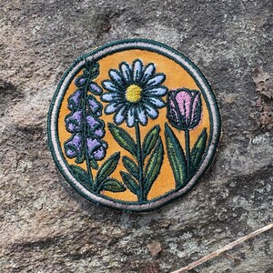 Pu&ograve; includere: Toppa ricamata con un design floreale. La toppa ha uno sfondo giallo e un bordo verde. Il design include una margherita, un tulipano e un gruppo di fiori viola. I fiori hanno foglie verdi.