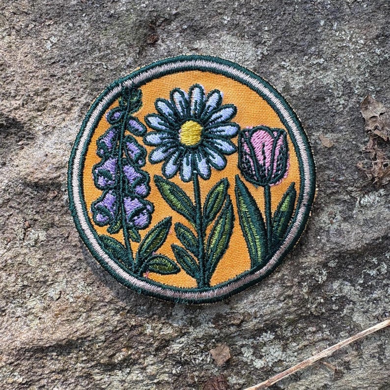 Pu&ograve; includere: Toppa ricamata con un design floreale. La toppa ha uno sfondo giallo e un bordo verde. Il design include una margherita, un tulipano e un gruppo di fiori viola. I fiori hanno foglie verdi.