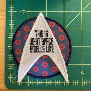 May include: Embroidered patch with a white and black design. The patch features a white arrowhead shape with the text "THIS IS WHAT SPACE SMELLS LIKE" in black. The patch is on a blue background with red circles.