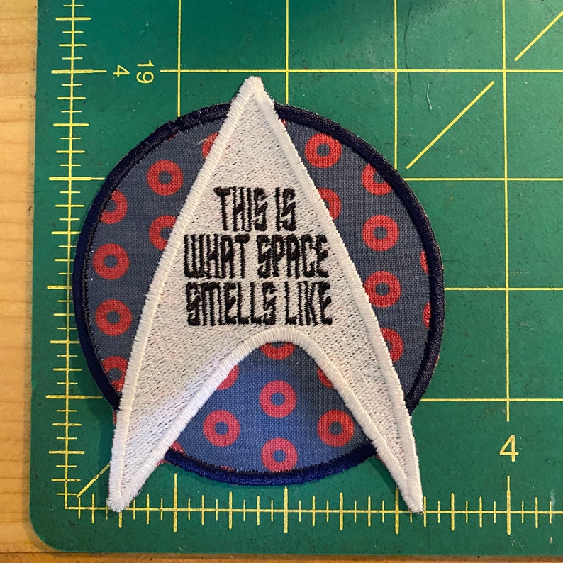 May include: Embroidered patch with a white and black design. The patch features a white arrowhead shape with the text "THIS IS WHAT SPACE SMELLS LIKE" in black. The patch is on a blue background with red circles.