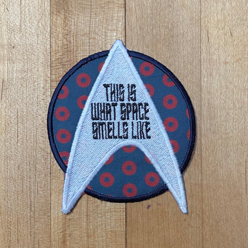 May include: Embroidered patch with a white and blue Starfleet insignia design. The patch is circular with a blue background and red donut-shaped patterns. Text reads "THIS IS WHAT SPACE SMELLS LIKE."