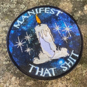 May include: Round patch with the text "MANIFEST THAT SHIT" in white lettering. The patch features a blue and black galaxy background with a white candle and orange flame. Decorative stars are also included.