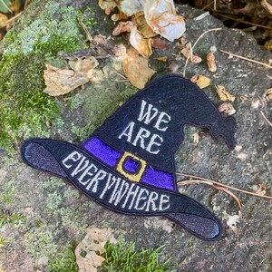 May include: Black witch hat patch with purple band and gold buckle. The words "WE ARE EVERYWHERE" are embroidered in white on the hat. The patch is set against a mossy, natural background.