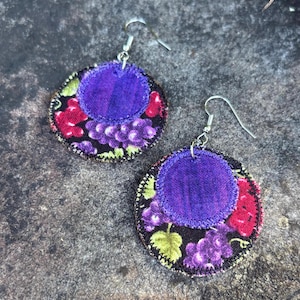 May include: A pair of handmade fabric earrings. Each earring features a circular design with a purple center and a grape pattern in shades of purple, red, and green on a black background. Silver-tone ear wires complete the design.