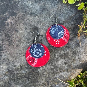 May include: A pair of handmade earrings featuring a red and navy blue paisley design. The earrings have a circular shape with a smaller navy blue circle in the center. The earrings are made of fabric and have silver-colored hooks.