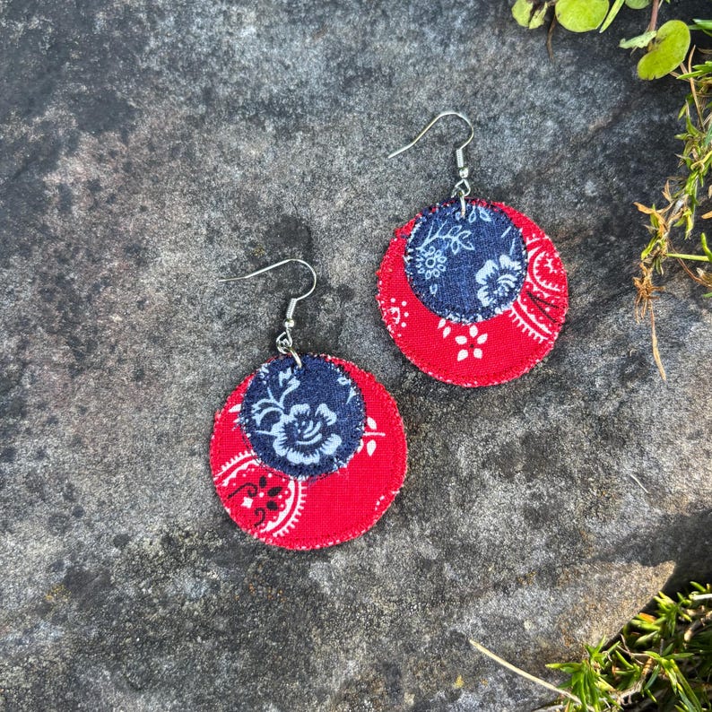 May include: A pair of handmade earrings featuring a red and navy blue paisley design. The earrings have a circular shape with a smaller navy blue circle in the center. The earrings are made of fabric and have silver-colored hooks.