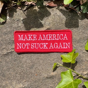 Może przedstawiać: Prostokątna czerwona naszywka z białym haftowanym napisem: "MAKE AMERICA NOT SUCK AGAIN". Naszywka umieszczona jest na teksturowanej kamiennej powierzchni, z zielonym bluszczem i liśćmi w tle.