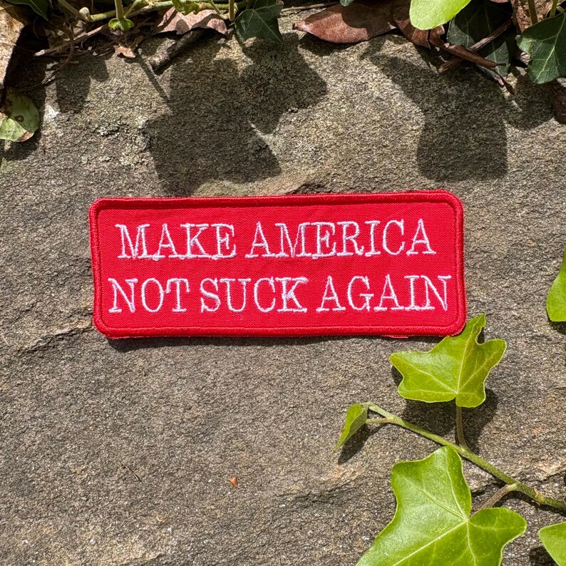 Może przedstawiać: Prostokątna czerwona naszywka z białym haftowanym napisem: "MAKE AMERICA NOT SUCK AGAIN". Naszywka umieszczona jest na teksturowanej kamiennej powierzchni, z zielonym bluszczem i liśćmi w tle.