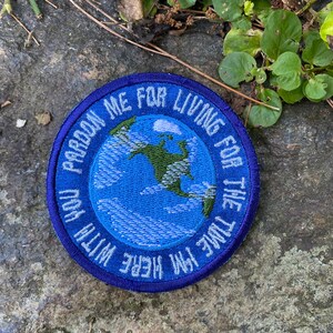 May include: A round, blue embroidered patch with the text "Pardon me for living for the time I'm here with you." The patch features a blue and green embroidered image of the Earth.