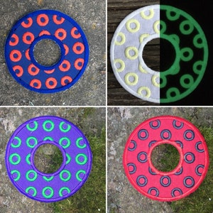 May include: Four colorful, circular coasters with a central hole. The coasters feature embroidered patterns of circles in various color combinations: blue with orange, white with yellow, purple with green, and red with blue. One coaster glows in the dark.