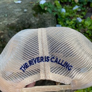 Może przedstawiać: Beżowa czapka typu trucker z siatkowym tyłem i zakrzywionym daszkiem. Tekst "THE RIVER IS CALLING" jest wyhaftowany w kolorze granatowym z tyłu. Czapka posiada zapięcie na zatrzask dla regulacji rozmiaru.