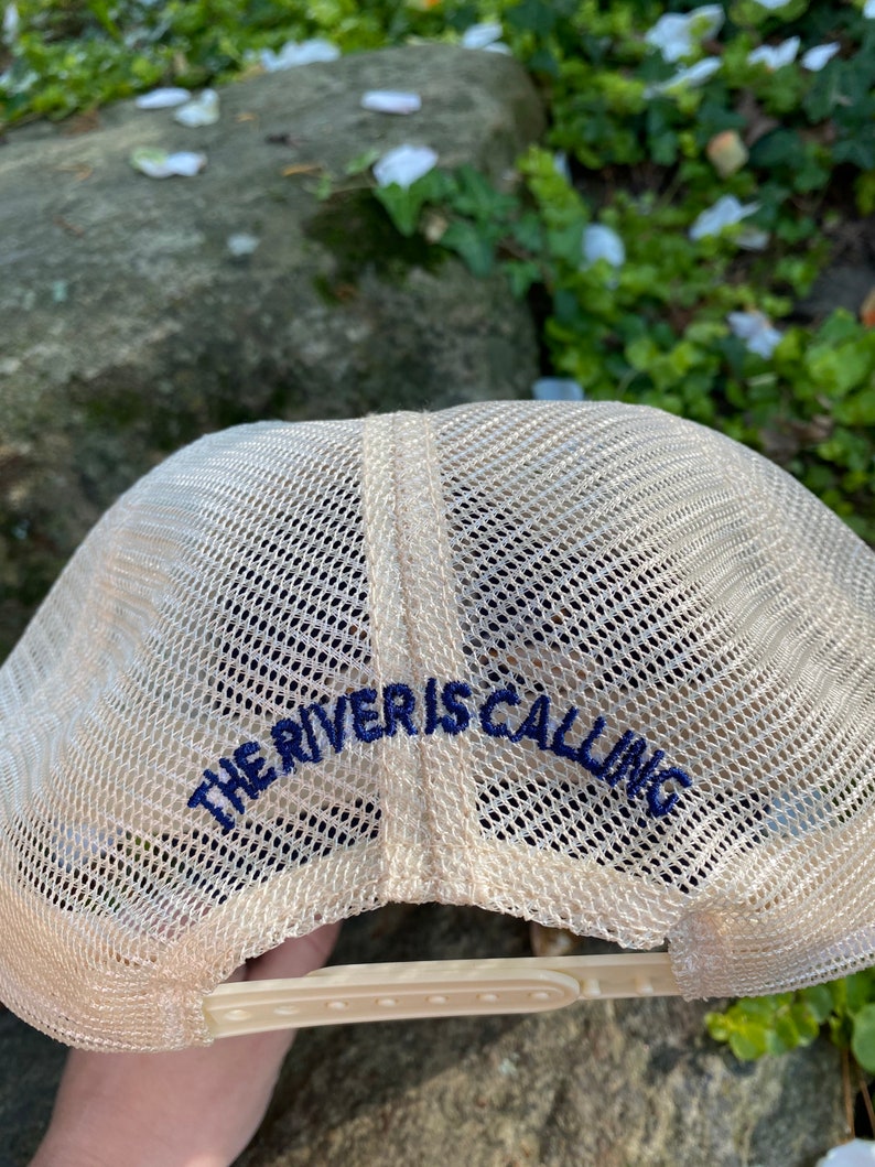 Może przedstawiać: Beżowa czapka typu trucker z siatkowym tyłem i zakrzywionym daszkiem. Tekst "THE RIVER IS CALLING" jest wyhaftowany w kolorze granatowym z tyłu. Czapka posiada zapięcie na zatrzask dla regulacji rozmiaru.
