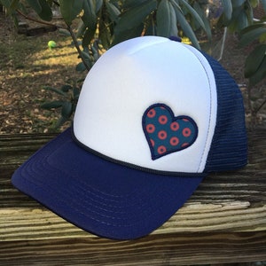 May include: A navy blue and white trucker hat with a heart-shaped patch. The heart is teal with red polka dots. The hat has a navy blue brim and mesh back, with a black cord detail.